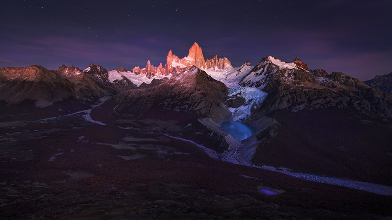 Jagged granite peaks illuminated in warm orange light at twilight, with a glacier and dark mountain valley below.