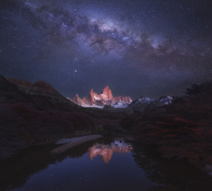 Illuminated mountain peaks reflected in still water beneath the Milky Way.