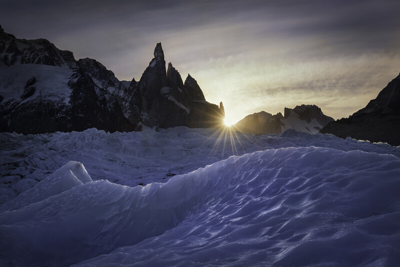 Snow-covered glacier landscape with dramatic rocky peaks silhouetted against a bright sun and cloudy sky.