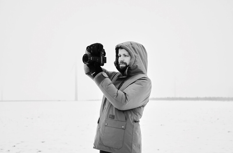 Bearded man in hoodie and blazer holding professional camera to his face against white background.