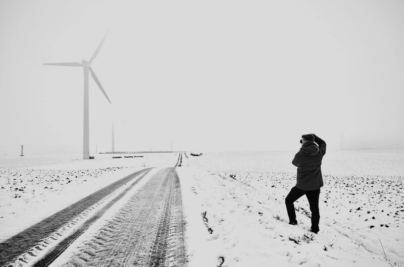 Person in dark winter coat standing in snowy field beside straight road with wind turbine in distance.