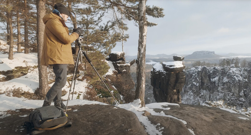 Photographer on a snowy overlook setting up a tripod-mounted camera overlooking a forested valley.
