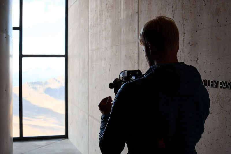 Photographer silhouetted against a bright window, holding a camera to their face while shooting a sunset landscape.