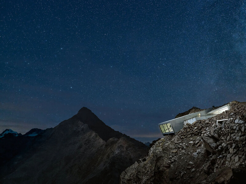 Illuminated structure perched on a rocky ridge at night with a silhouetted mountain peak and starry sky beyond.