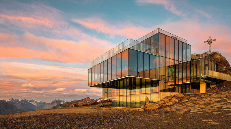 Modern glass and steel structure with overhanging design perched on rocky alpine terrain at sunset.