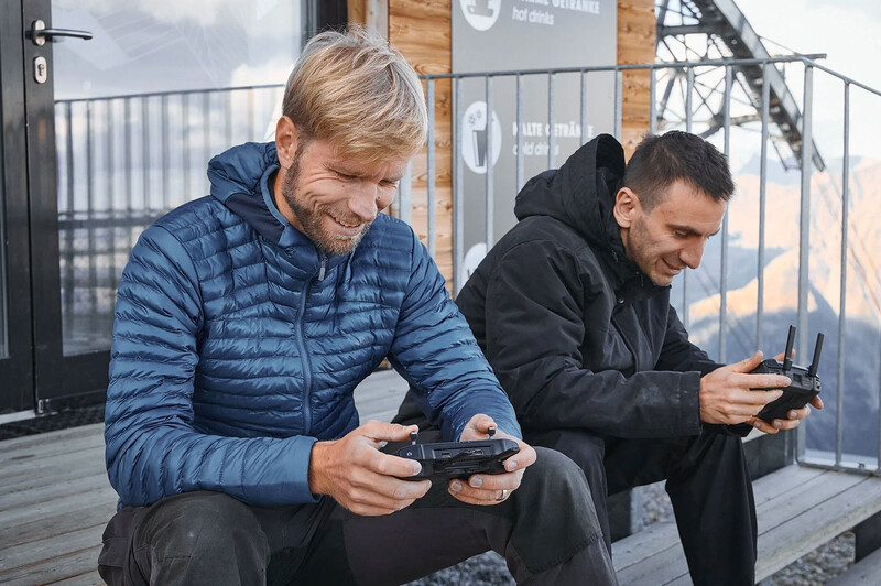 Two men holding drone remote controllers on an elevated wooden deck overlooking a city skyline.