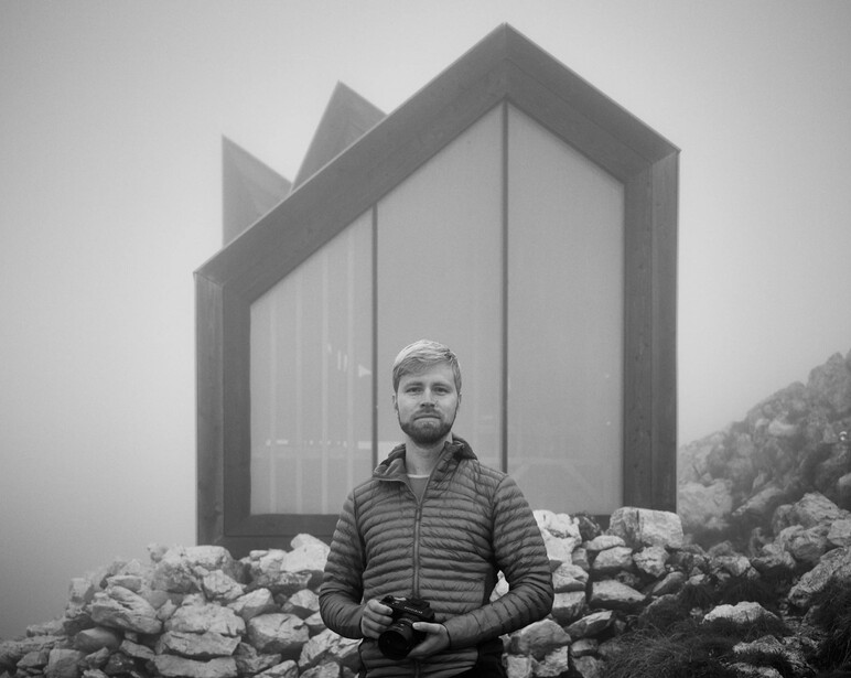 Man in striped jacket stands before a modernist house structure with large windows, surrounded by rocks in misty conditions.