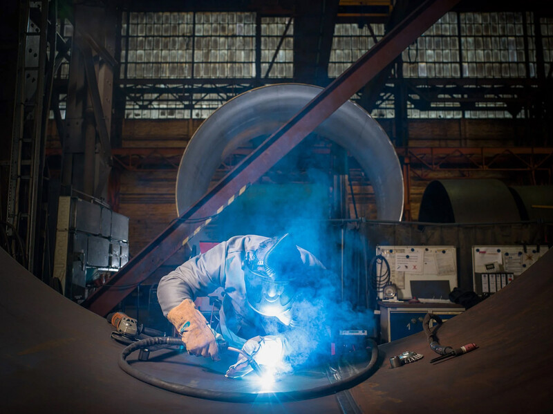 Welder performing arc welding inside a transparent protective dome with bright blue light illuminating the workspace.