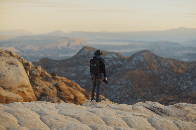 Solo photographer stands on sandstone ledge overlooking snowy Utah mountain range with camera and hiking backpack.