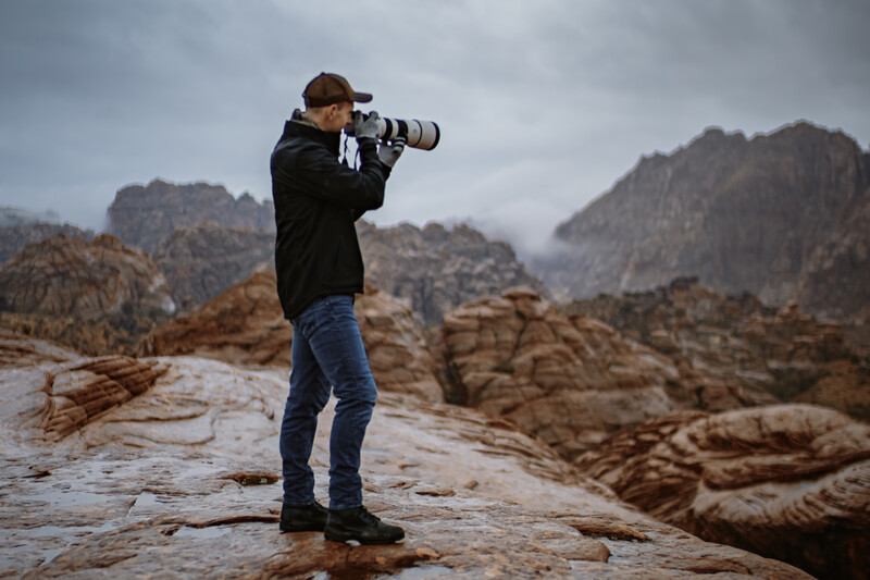 Photographer in Snow Canyon capturing brand imagery on wet red rocks with dramatic mountain backdrop and stormy sky.