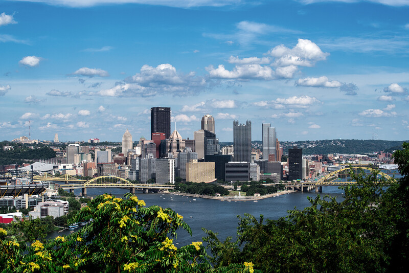 Pittsburgh skyline viewed from elevated vantage point with river in foreground and blue sky with white clouds.