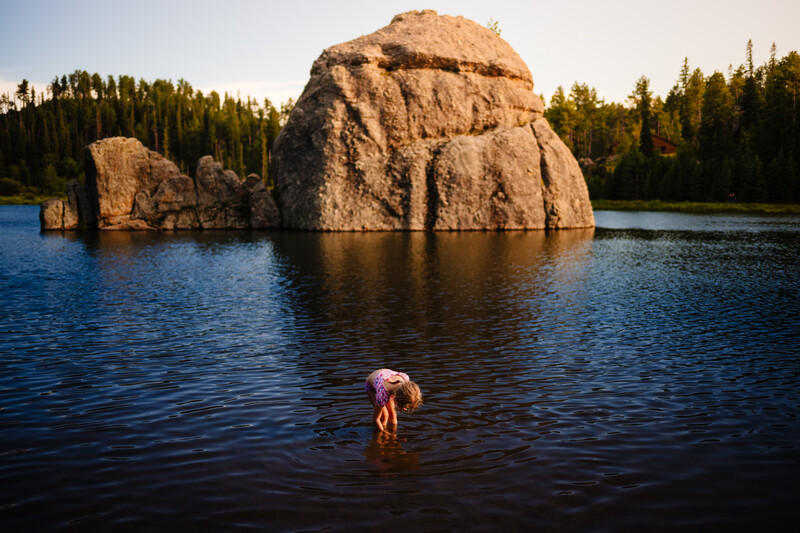 Person wading in calm lake water with massive granite boulder formation and coniferous forest in background.