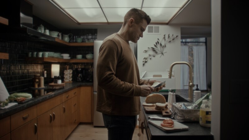 Man in brown cardigan stands at kitchen sink looking at smartphone while preparing food.