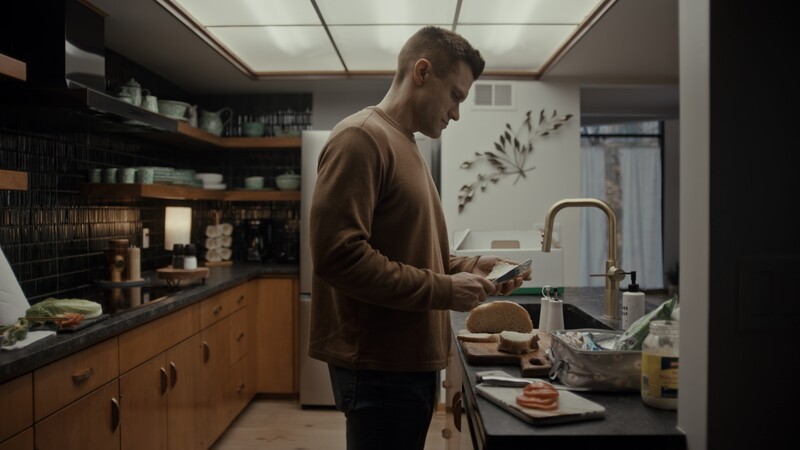 Man in brown sweater looking at phone while standing at kitchen sink with breakfast items.