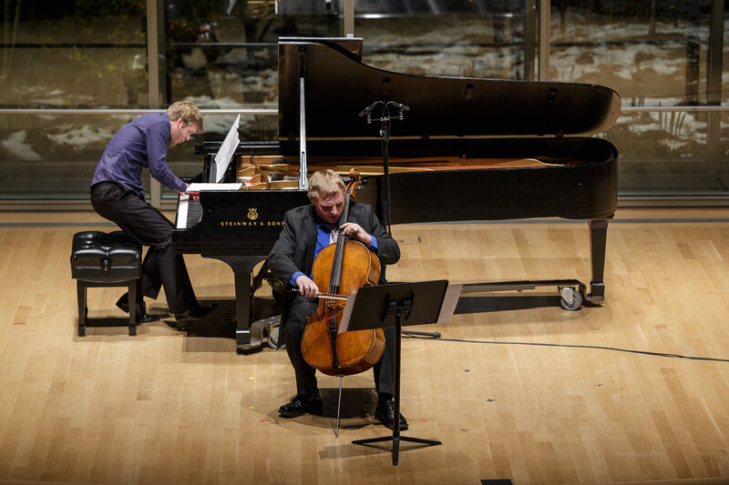 Pianist and cellist performing together on a concert hall stage with a grand piano.