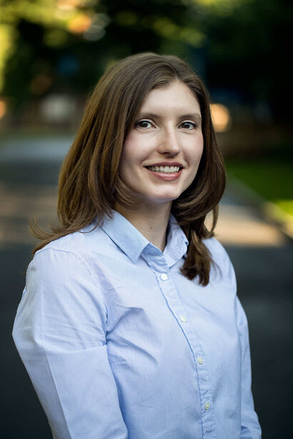 Woman with long brown hair smiling at camera wearing light blue button-up shirt.