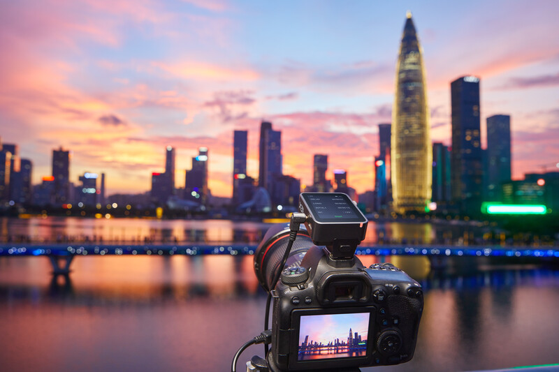 Camera on tripod displaying cityscape on LCD screen with illuminated skyscrapers and sunset sky reflected in water behind it.