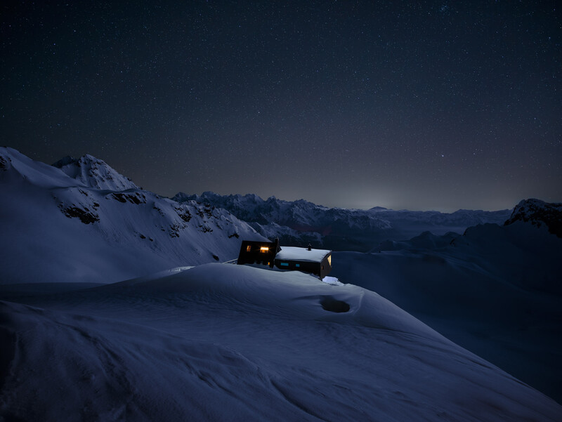 Illuminated cabin on a snowy mountain ridge under starlit night sky with distant peaks.