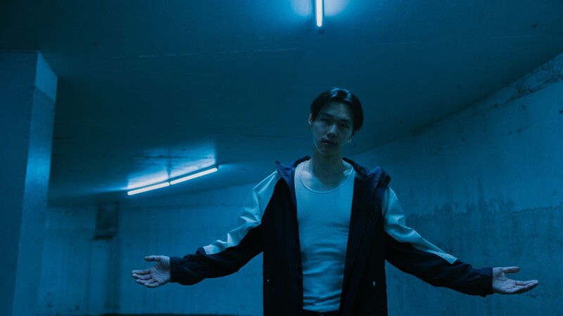 Young man in dark jacket and white shirt stands in fluorescent-lit hallway with arms outstretched.