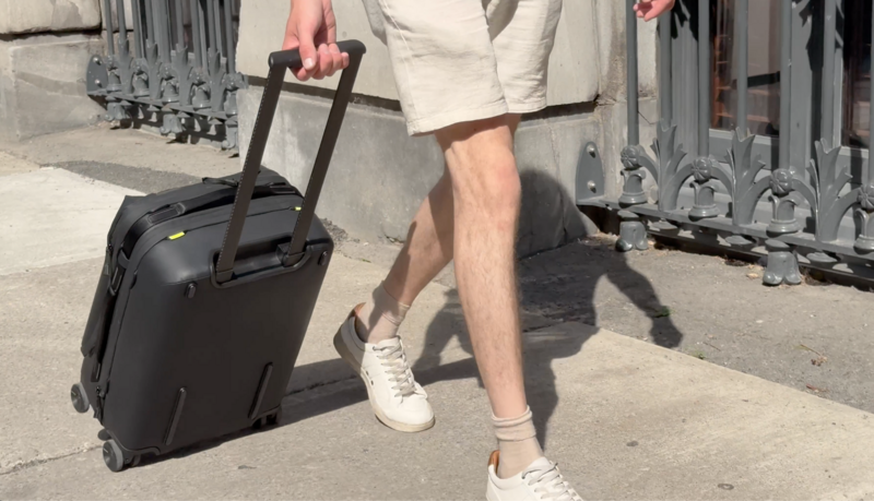 Person in cream jacket and white sneakers pulling black rolling luggage on a sunny street.