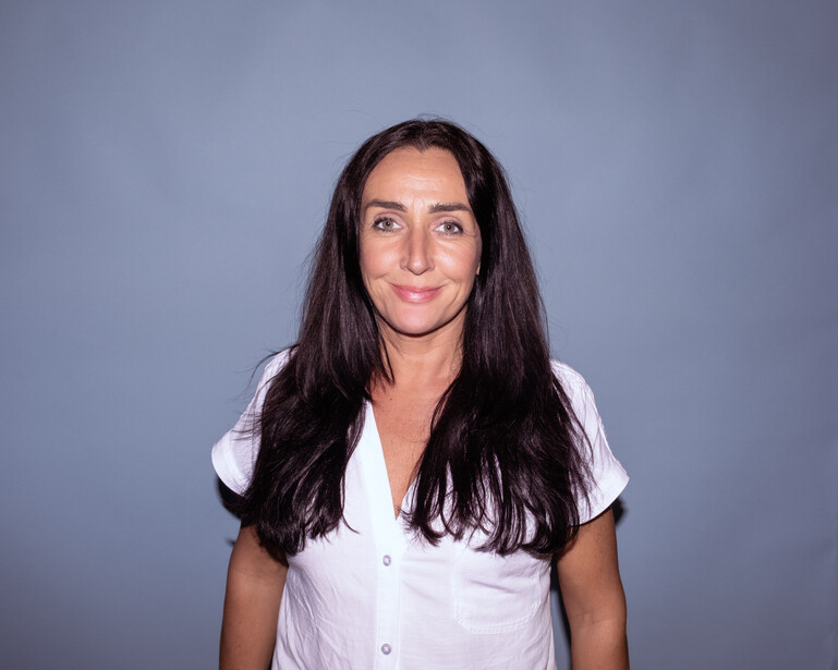 Woman with long dark hair wearing a white button-up shirt against a neutral gray background.