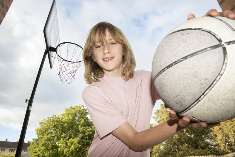 Young person holding a basketball outdoors near a basketball hoop, wearing a pink t-shirt.