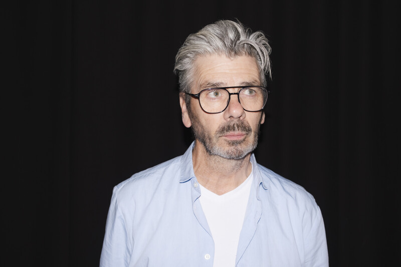 Middle-aged man with gray hair and glasses wearing a light blue shirt against a black background.
