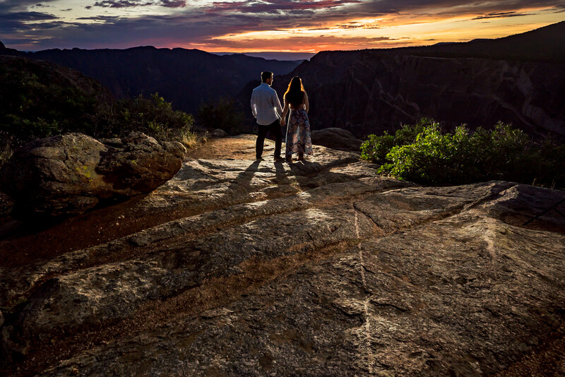 Two people standing on a rocky overlook at sunset above a vast canyon landscape.
