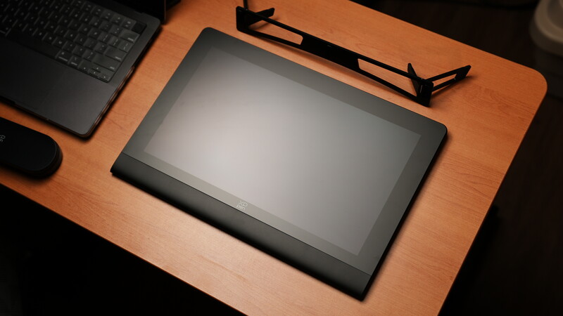 Tablet with blank screen on wooden desk next to laptop, mouse, and monitor stand.