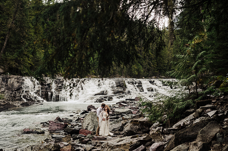 Bride in white wedding dress standing on rocky riverbank with cascading waterfall and dense forest backdrop.