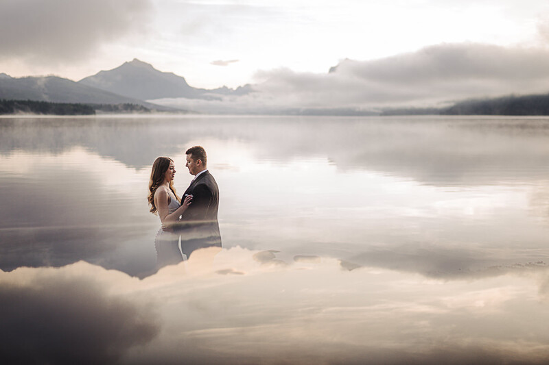 Couple standing in shallow lake water with misty mountains reflected in calm surface.