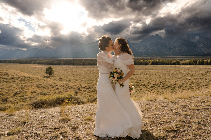 Two brides kiss in an open field with dramatic storm clouds and mountains in the background.