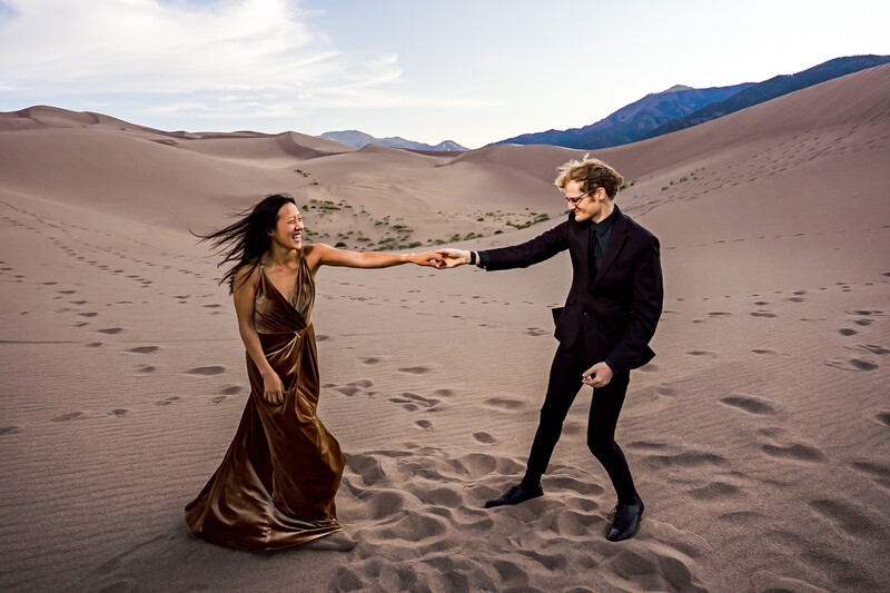 Couple holding hands on vast sand dune with mountains under cloudy sky.