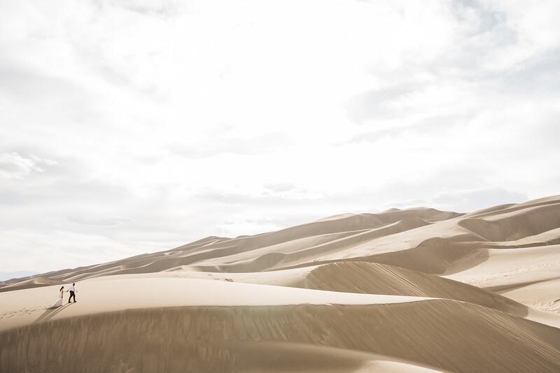 Two hikers walking across vast sand dunes under overcast sky.