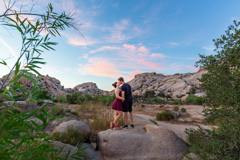 Couple embracing on a large boulder in a desert landscape with granite rock formations and scattered vegetation.