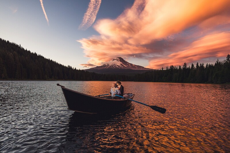 Person in rowboat on calm lake at sunset with snow-capped mountain and forest in background.