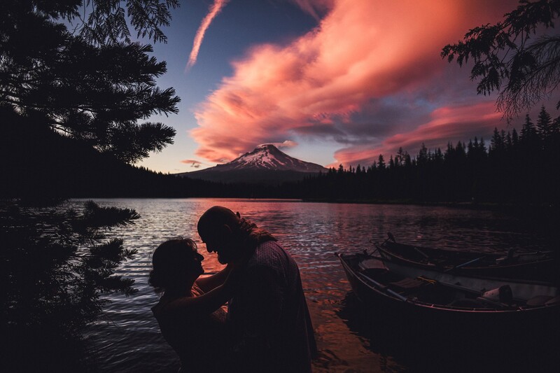 Two silhouetted figures embrace on a lakeshore at sunset, with a snow-capped mountain and dramatic orange-pink clouds reflected in the water.