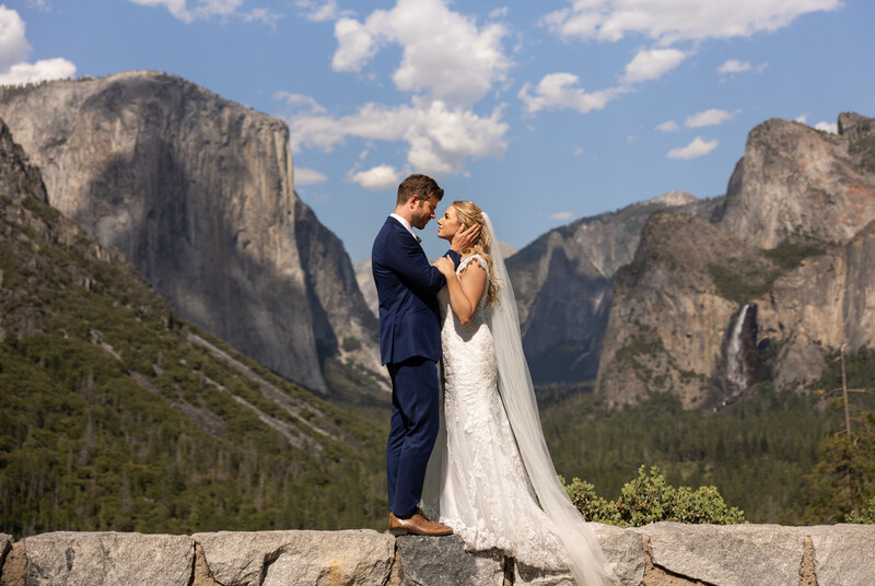 Bride and groom embrace on a stone overlook with dramatic granite cliffs and forested valley in background.