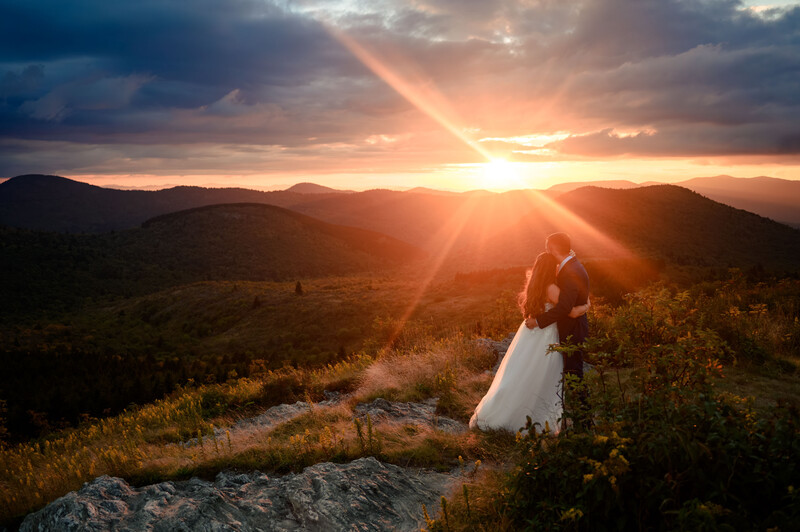 Bride and groom standing on a rocky hilltop during golden hour sunset, mountains visible in distant background.