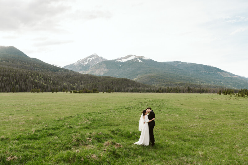 Couple in wedding attire embracing in a vast green meadow with snow-capped mountains in the background.