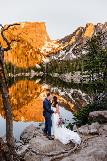 Bride and groom standing by an alpine lake with golden cliffs reflected in still water at sunset.
