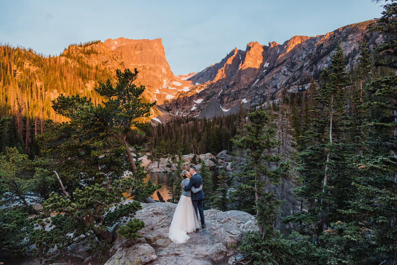 Couple in wedding attire standing on a rocky trail with alpine mountains and forest in golden hour light.