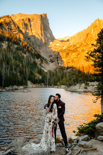 Couple in formal wear standing on rocks by a mountain lake at golden hour.