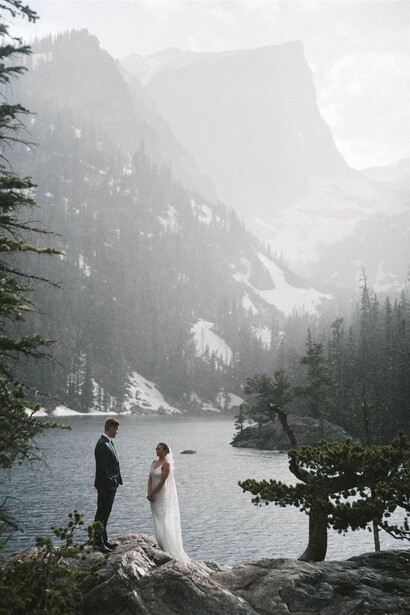 Couple standing on a rocky shore facing a misty alpine lake surrounded by steep mountain cliffs and evergreen trees.