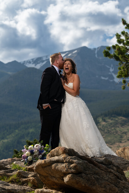 Bride and groom standing on a large rock with snow-capped mountains in the background.