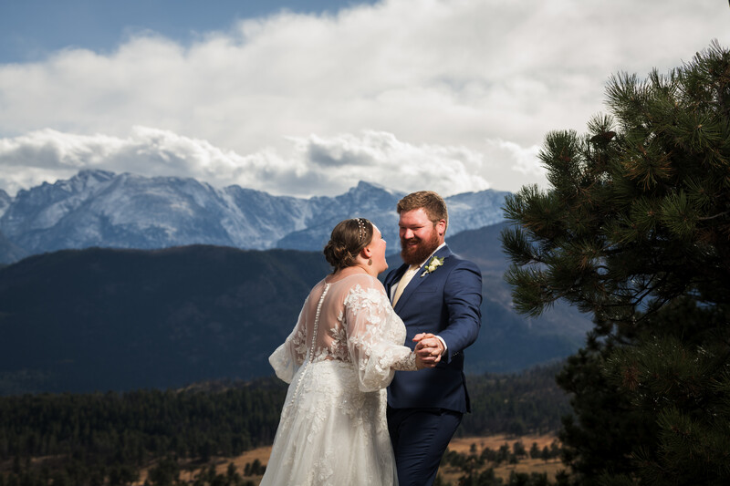 Bride and groom facing each other with snow-capped mountains and forested valley in background.