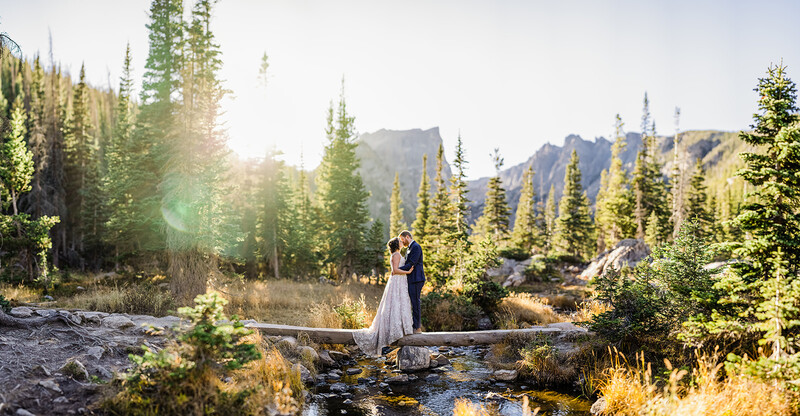 Couple standing on a fallen log over a stream in a coniferous forest with mountains in the background.