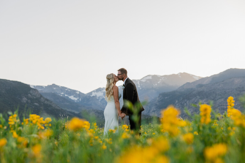 Bride and groom kissing in a field of yellow wildflowers with mountains in the background.
