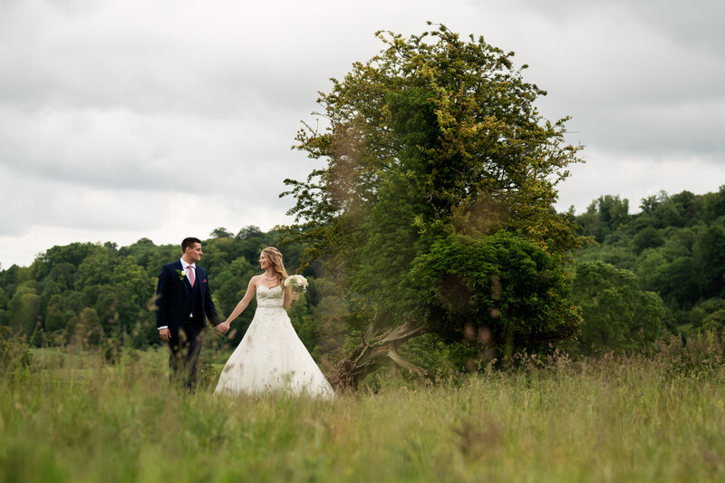 Bride and groom walking hand-in-hand through tall grass with trees and overcast sky in background.