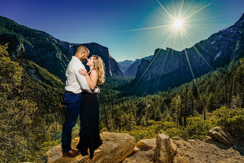 Couple kissing on a rocky overlook with a sunlit mountain valley and forested slopes in the background.